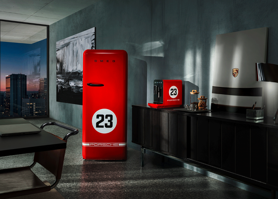 A modern, stylish interior with a large window revealing a cityscape at dusk. The room features sleek dark furniture and minimalist decor. Prominently displayed are a red SMEG refrigerator and a red coffee machine, each marked with a white circle containing the number "23" and "PORSCHE" logo. The refrigerator has the brand name "SMEG" in bold letters at the top. Behind it, a black and white abstract painting adds contrast to the scene. The design emphasizes bold colors, clean lines, and strong visual contrast, ensuring legibility and accessibility for users with visual impairments.